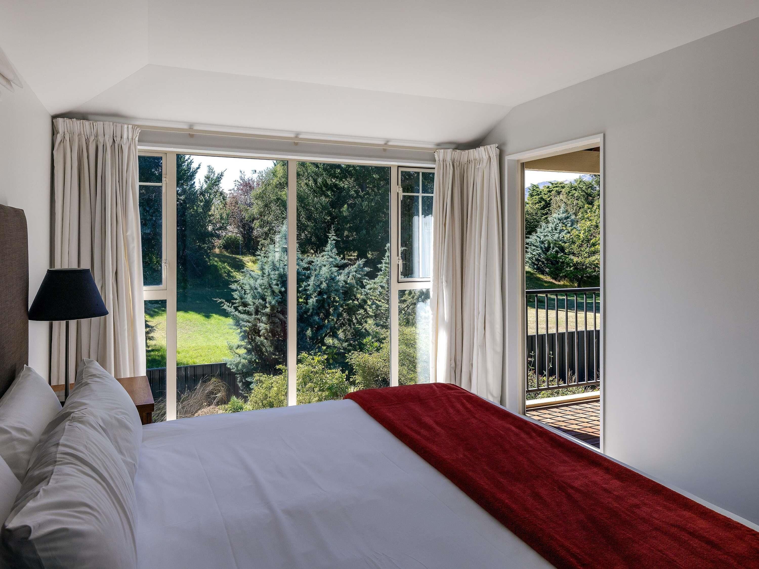 A bedroom with a large bed, a nightstand, a lamp, and a view of a garden through a sliding glass door.