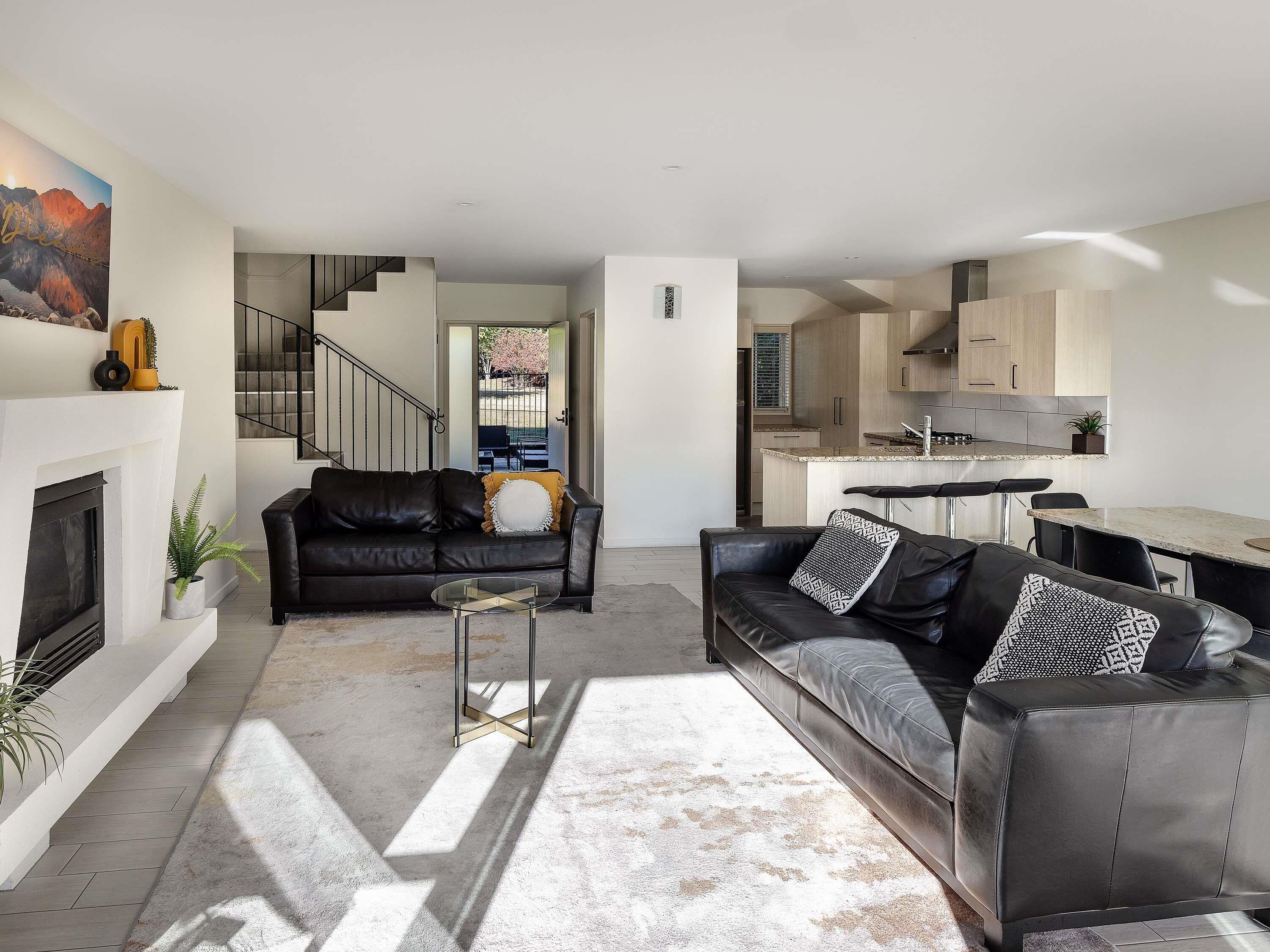 A modern living room with a fireplace, a black leather sofa, a glass coffee table, and a staircase leading to a kitchen.