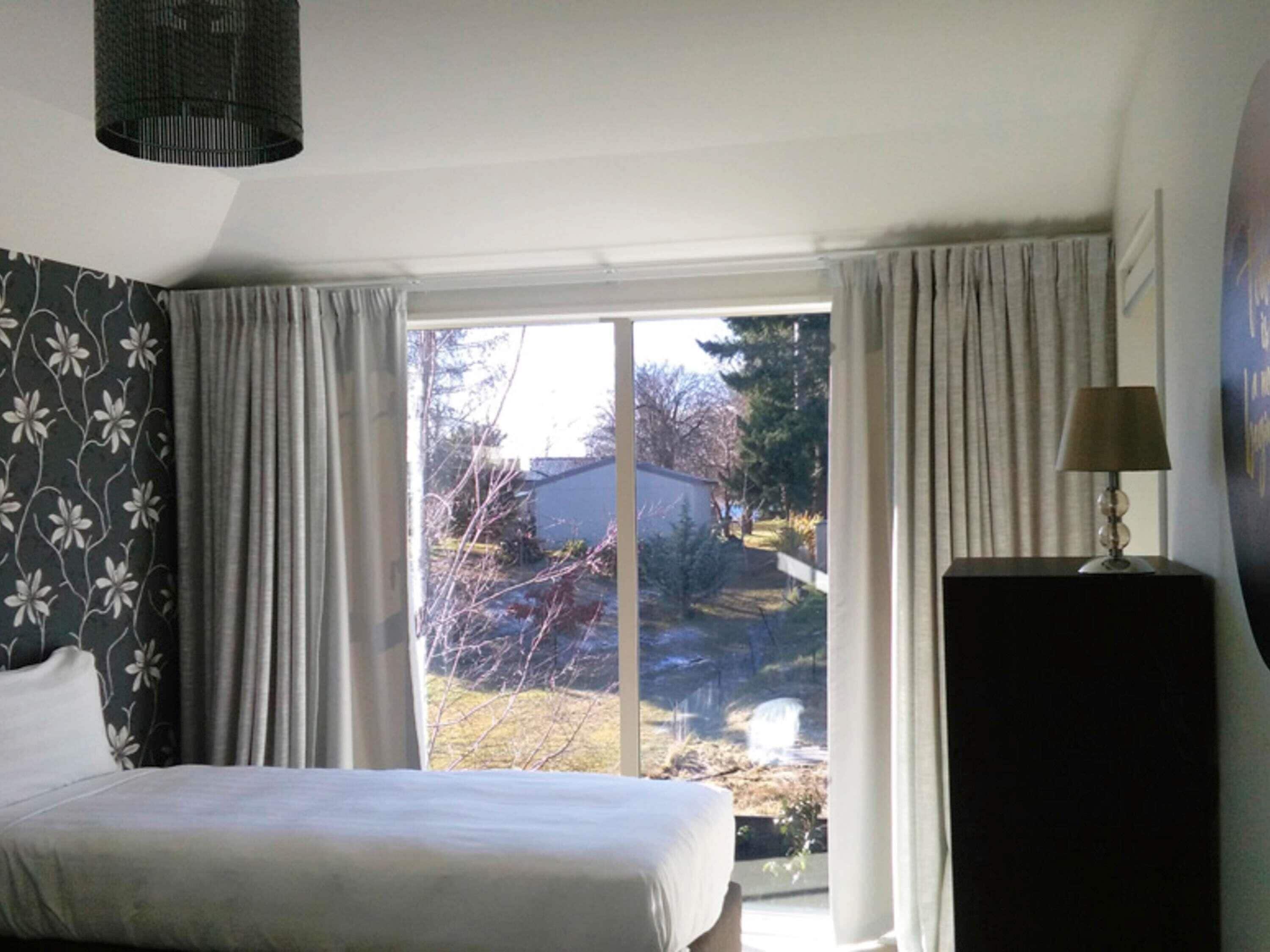 A bedroom with a bed, a window with curtains, a lamp, and a view of the outdoors.