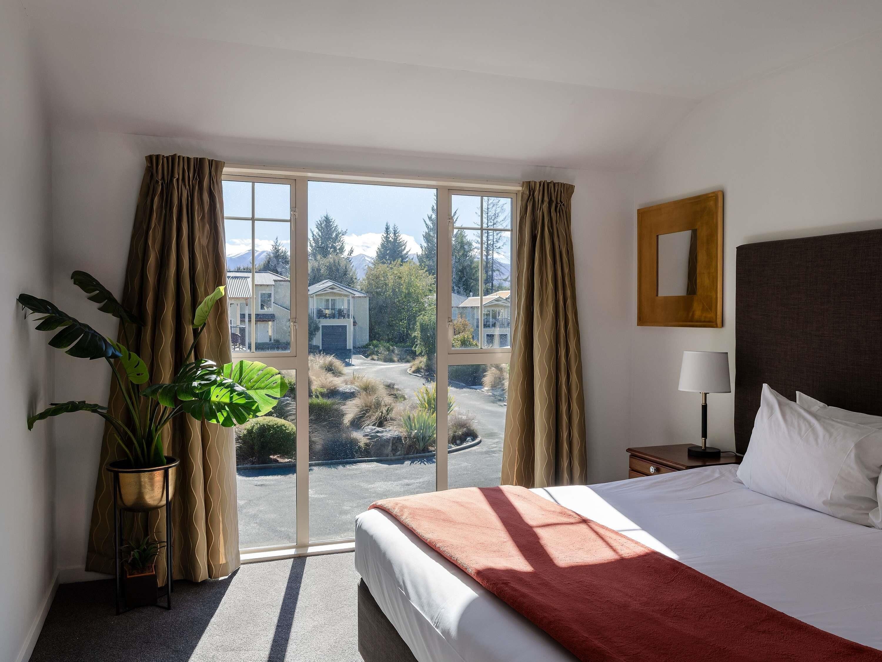 Une chambre d’hôtel avec un lit, une table de chevet, une lampe, une fenêtre avec des rideaux et une plante en pot.