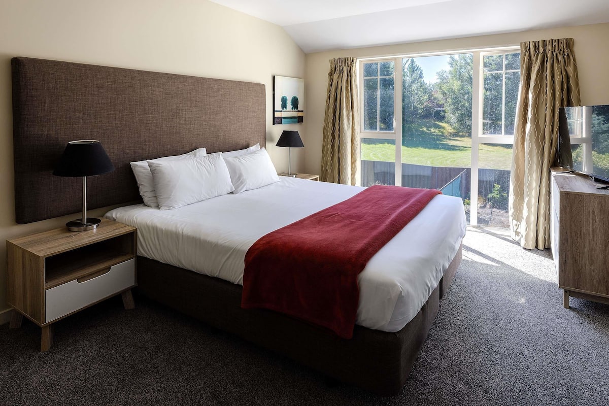 A hotel room with a large bed, a nightstand, a TV, and a window with curtains.