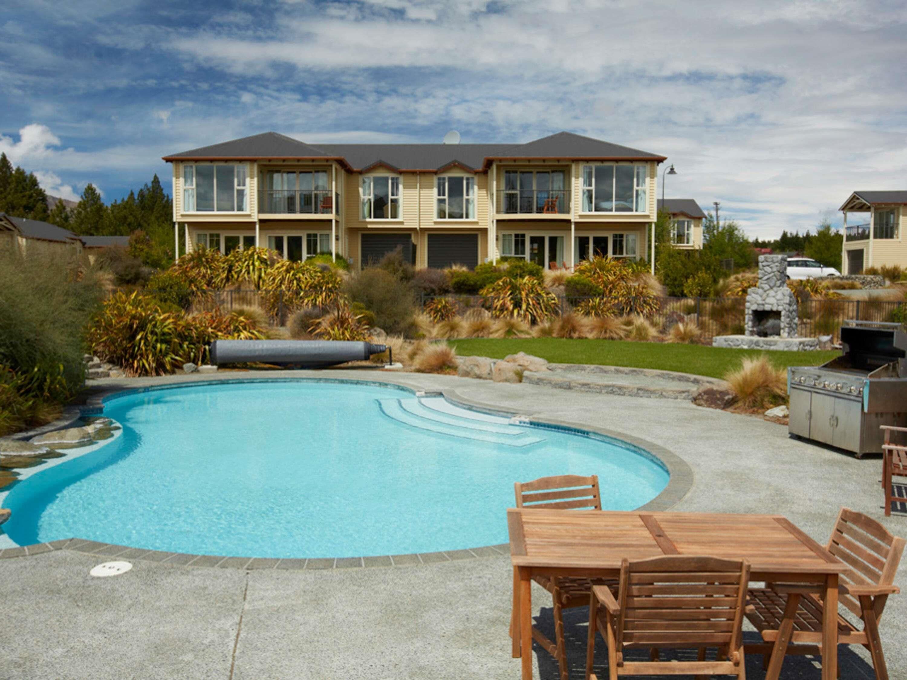 A swimming pool with a wooden deck and chairs, surrounded by a landscaped garden and a multi-story building with balconies.