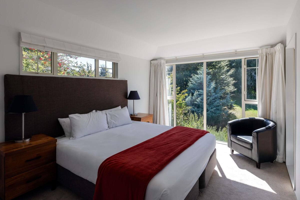 A bedroom with a large bed, two bedside tables, a chair, and a view of trees through large windows.