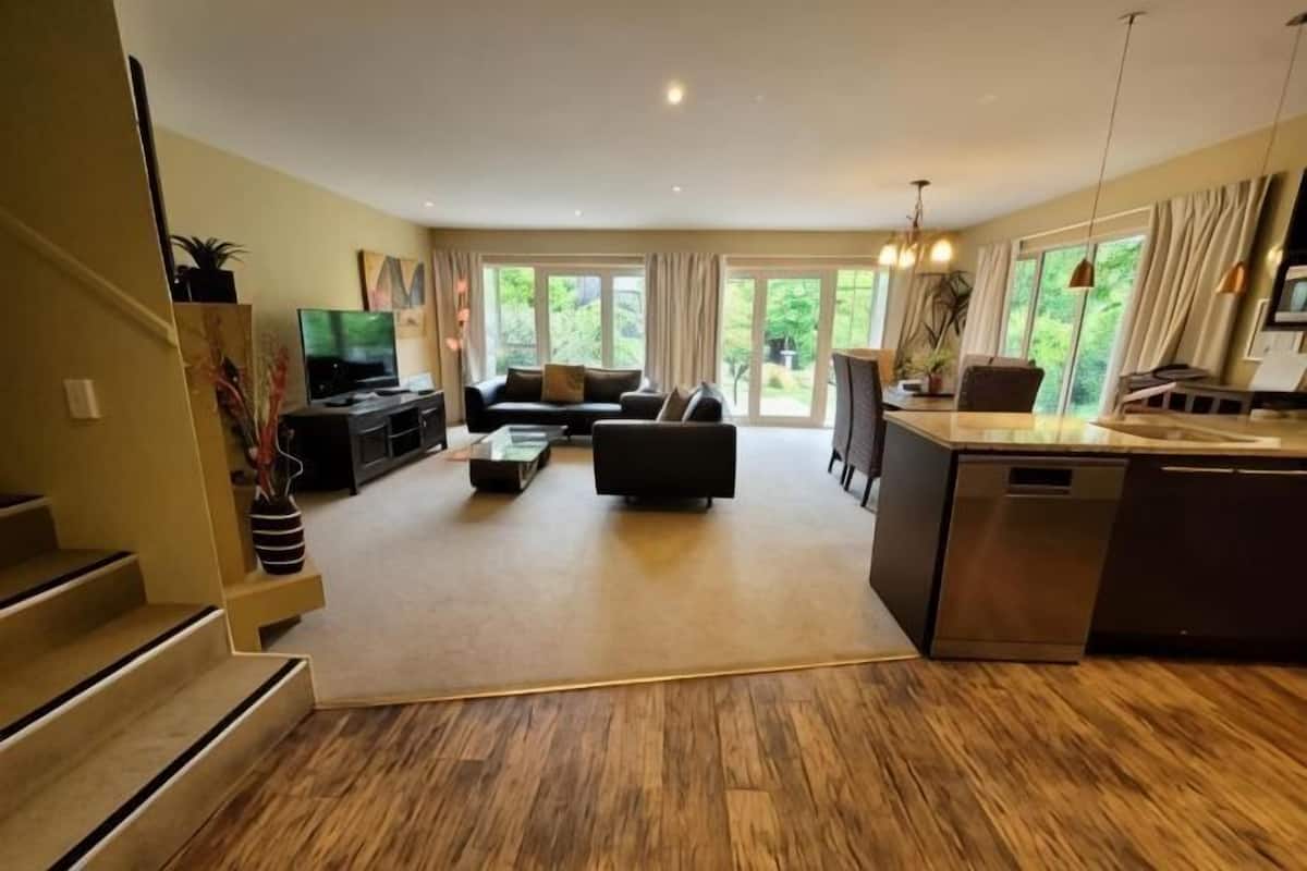 A modern living room with a sofa, coffee table, and TV. There is a kitchen area with a sink and counter.