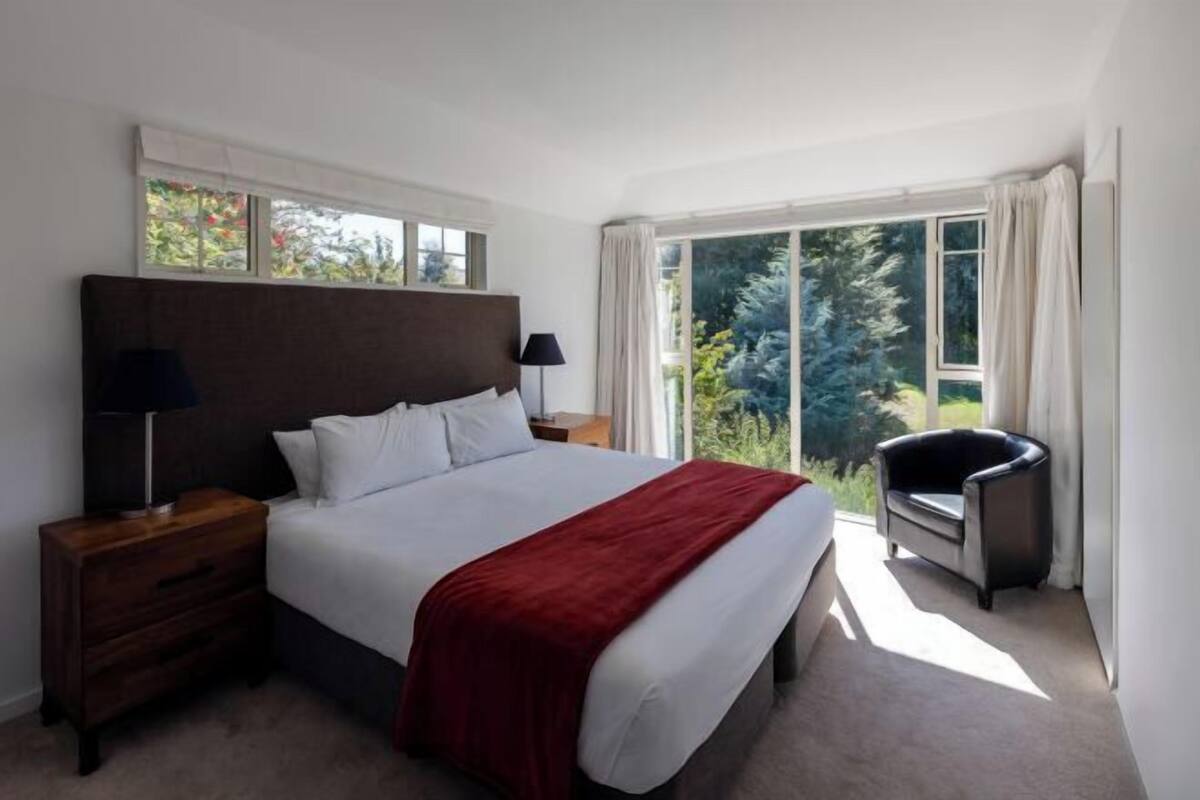 A bedroom with a large bed, a nightstand, a chair, and a window with a view of greenery.
