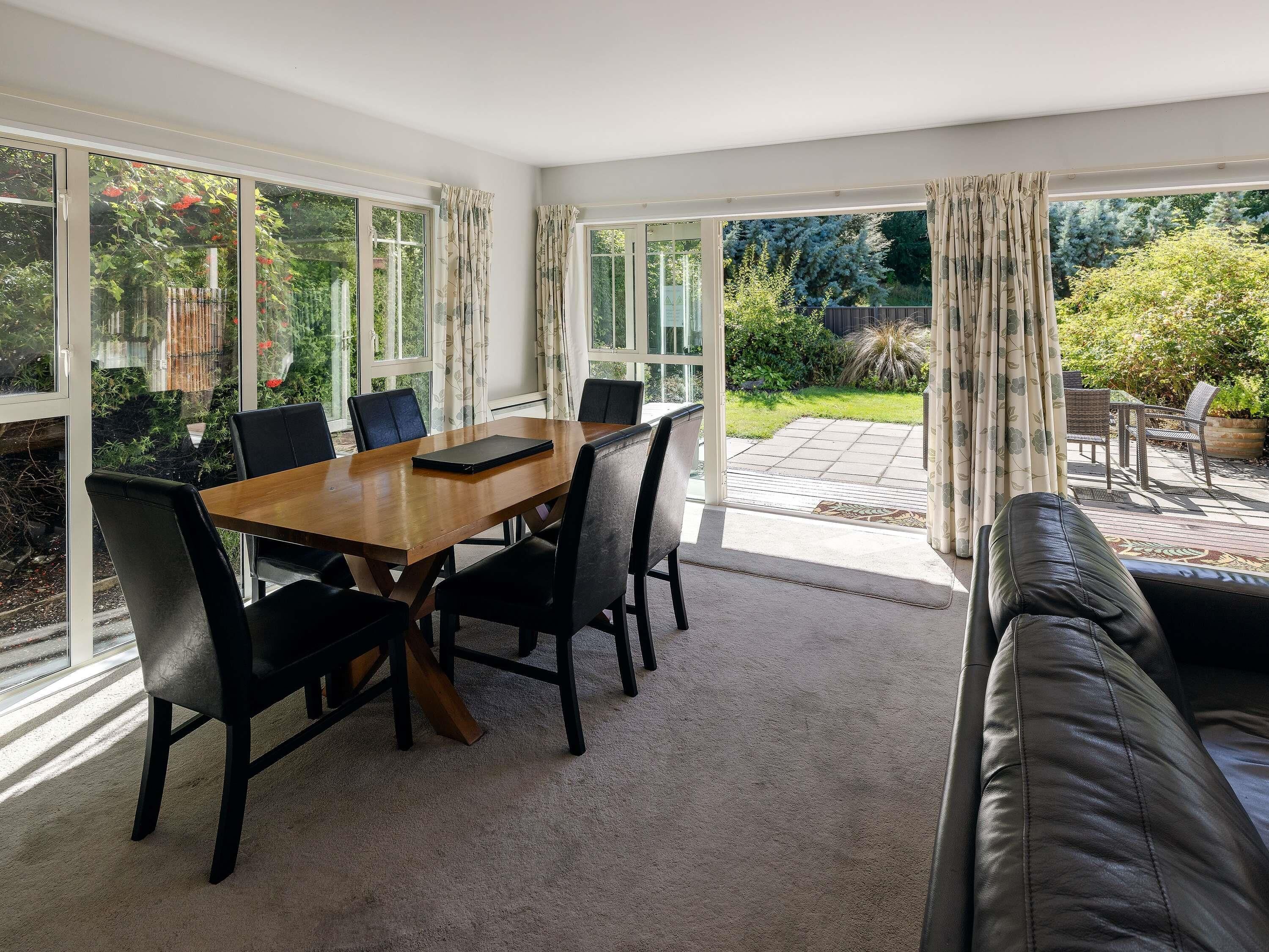 A room with a wooden dining table, black chairs, a leather sofa, and large windows with curtains.