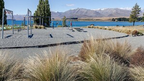 Un parque infantil con columpios y un tobogán, una zona de grava y un lago con montañas al fondo.