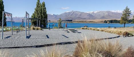 A playground with swings and a slide, a gravel area, and a lake with mountains in the background.