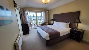 A hotel room with a large bed, a nightstand with a lamp, a dresser, and a view of the outside balcony.