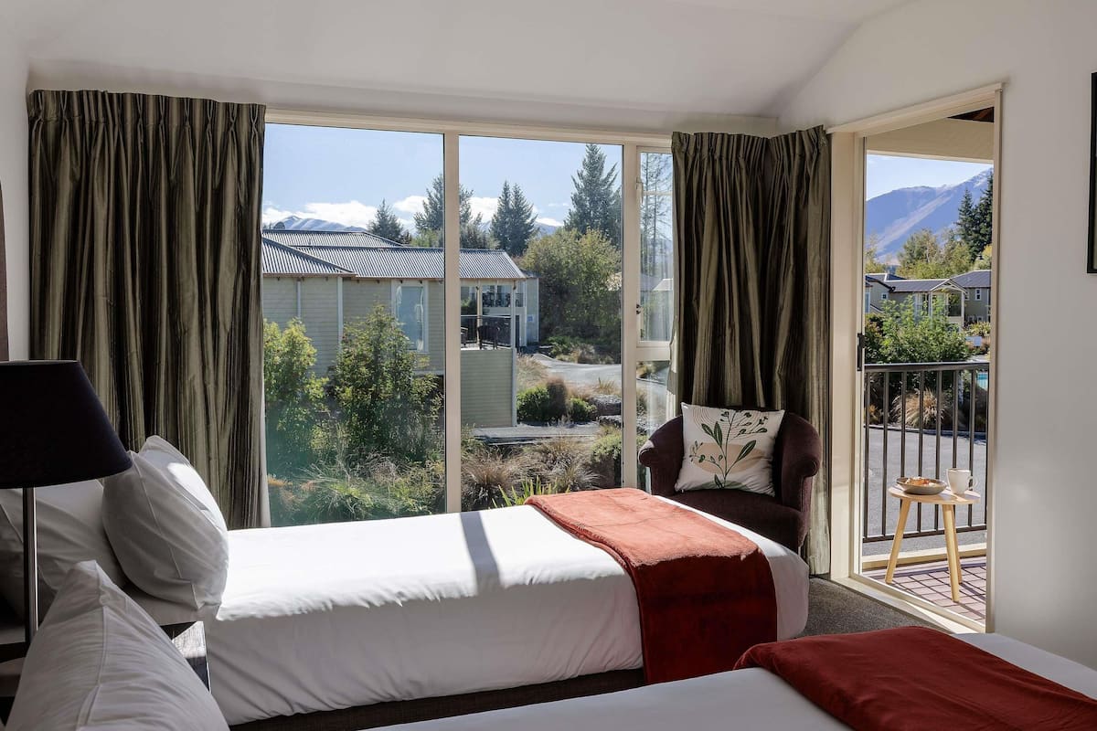 A hotel room with two beds, a chair, a balcony with a view of houses and mountains.