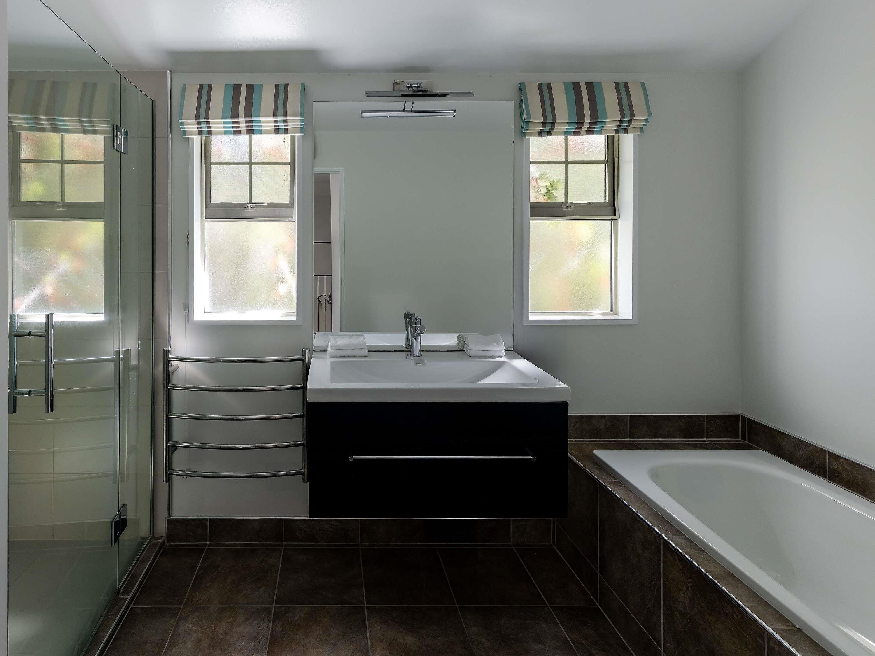 Une salle de bain avec une douche à parois de verre, un lavabo blanc avec un miroir, deux fenêtres ornées de rideaux à rayures et une baignoire.