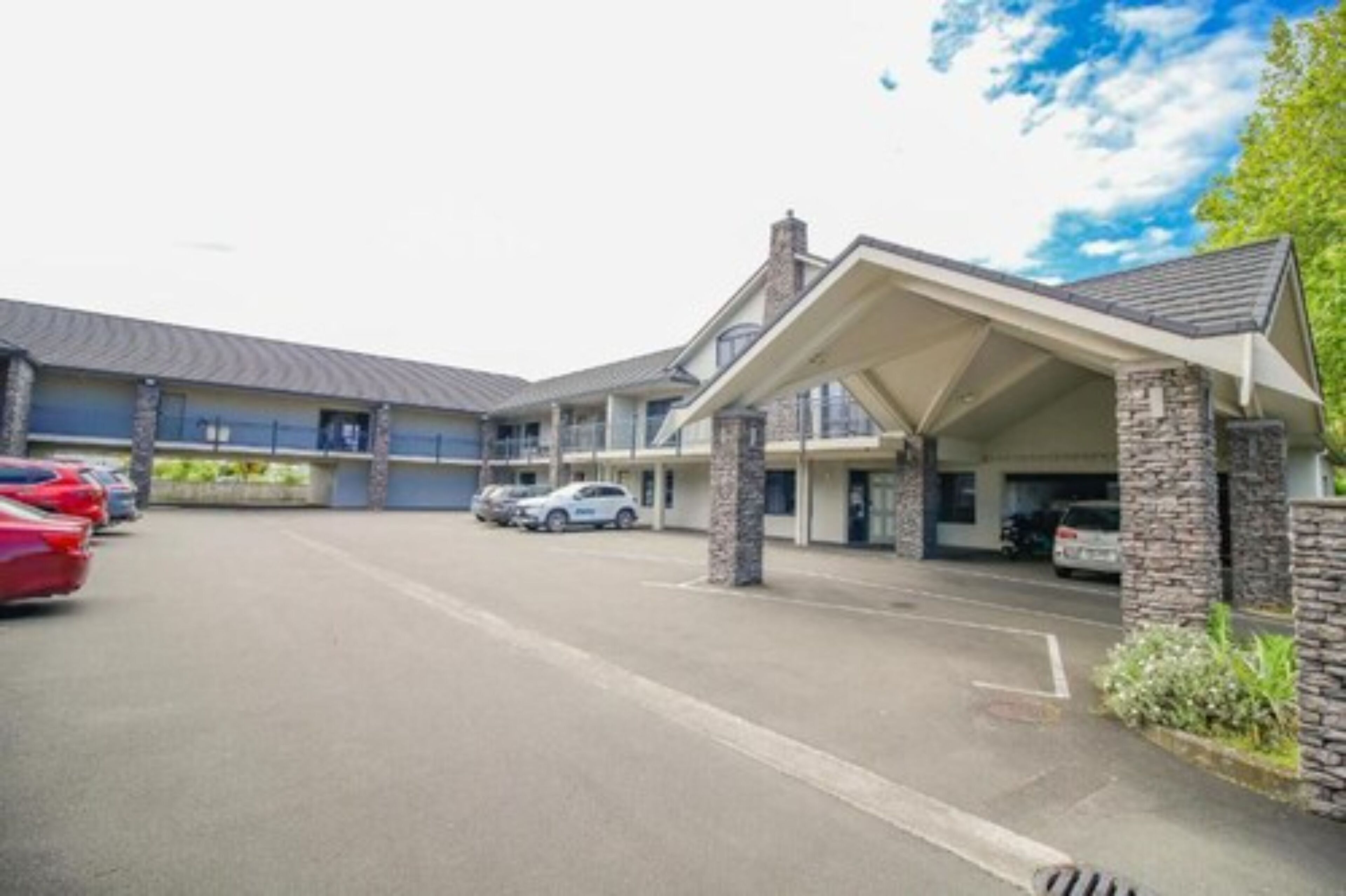 A motel with a covered entrance, a parking lot with several cars, and a building with a stone facade.