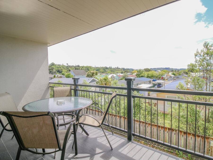 A balcony with a glass table and chairs, offering a view of a residential area with houses and greenery.