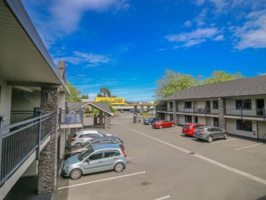 Un estacionamiento con varios coches aparcados frente a un motel que tiene balcón y una pequeña zona cubierta.