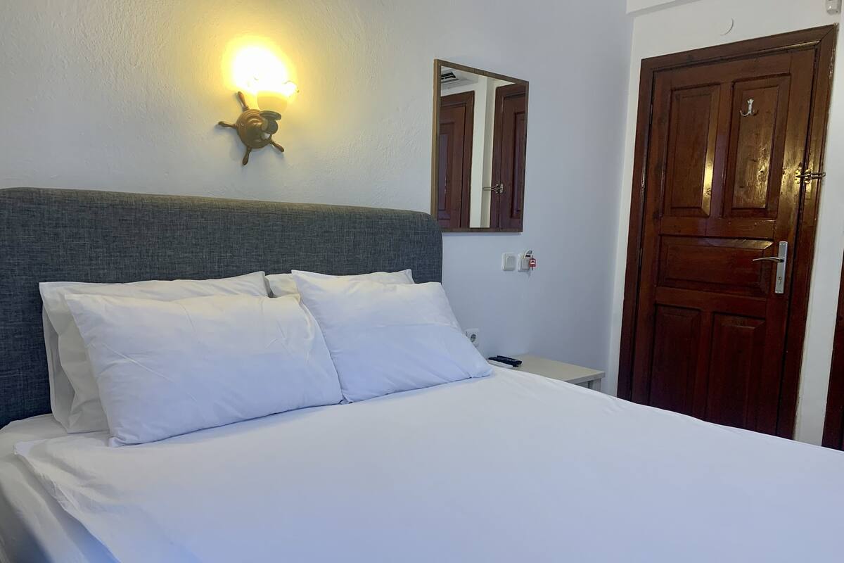 A neatly made bed with white linens, a wall-mounted light fixture, a mirror, and a wooden door.