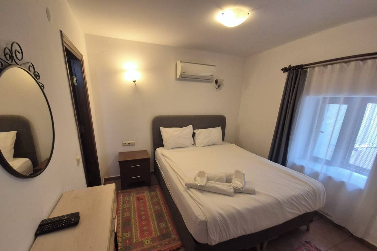 A bedroom with a bed, a bedside table, a mirror, an air conditioning unit, and a window with curtains.