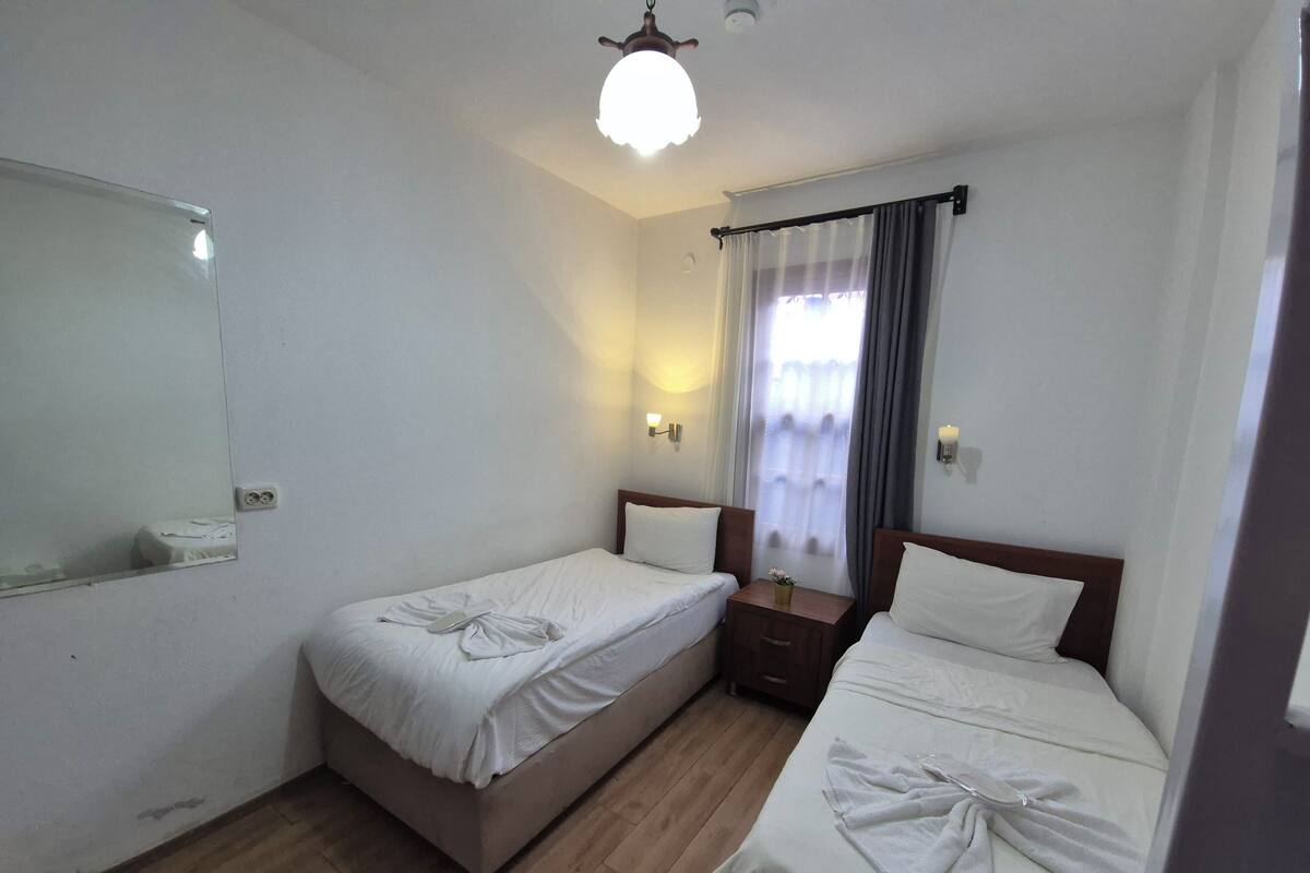 A hotel room with two single beds, a wooden floor, a mirror, a bedside table with a lamp, and a window with curtains.