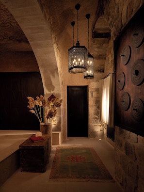 A dimly lit hallway with stone walls, a wooden door, and a decorative wall with circular patterns. There is a wooden bench with a vase of dried flowers and a red book.