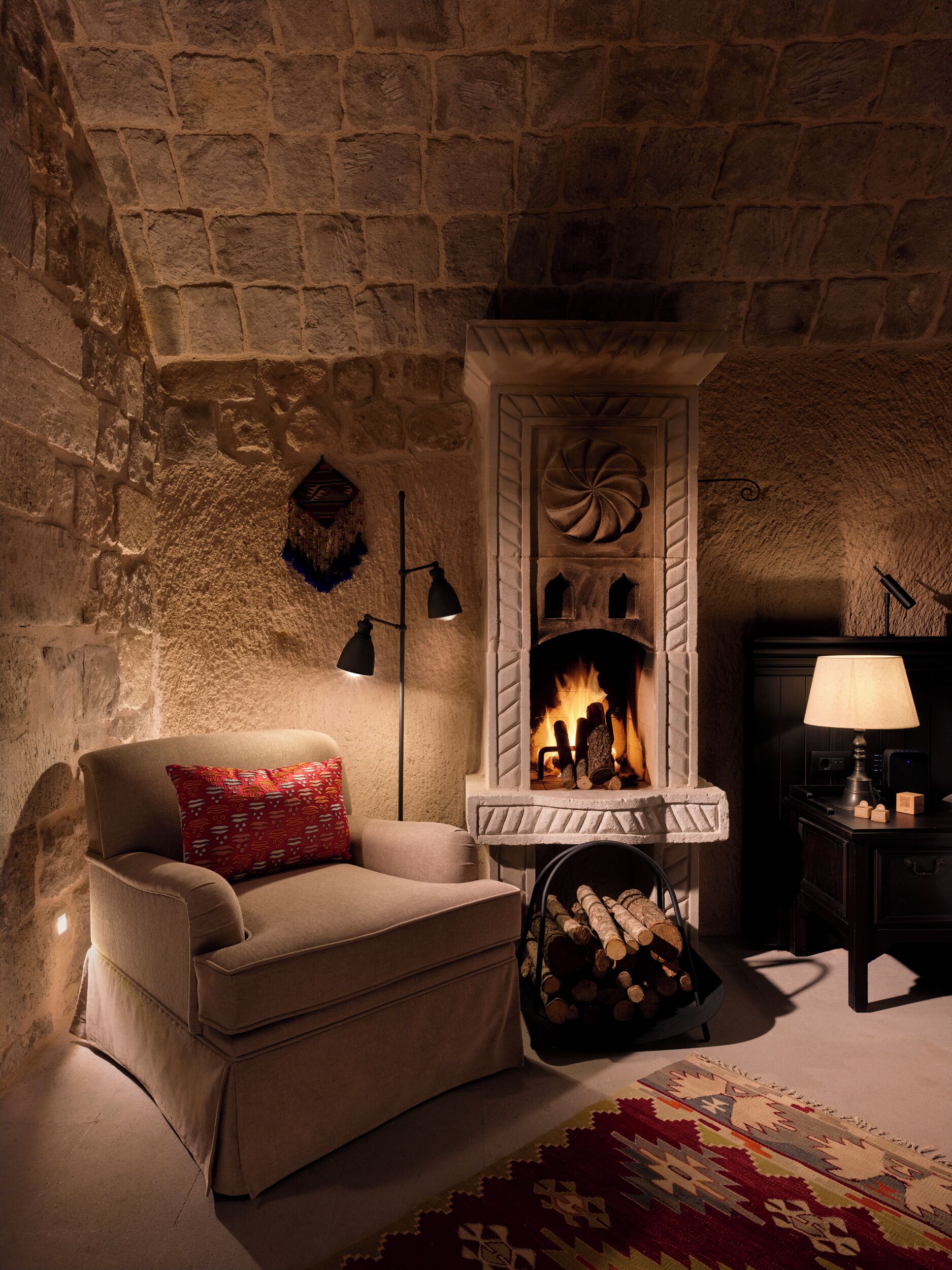 Una habitación acogedora con chimenea de piedra, un sofá con un cojín rojo y una alfombra estampada.