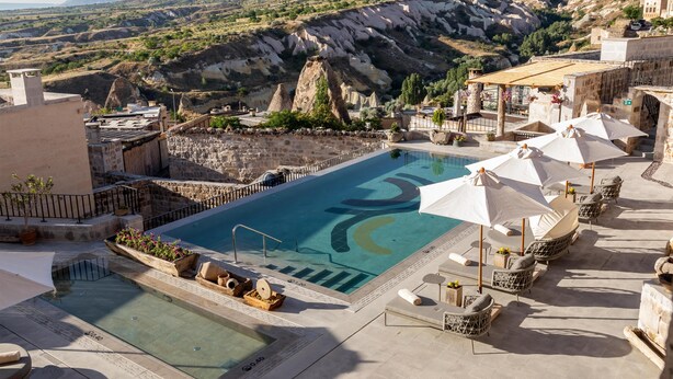 Un área de piscina con sillones de descanso y sombrillas, rodeada de muros de piedra y un paisaje montañoso.