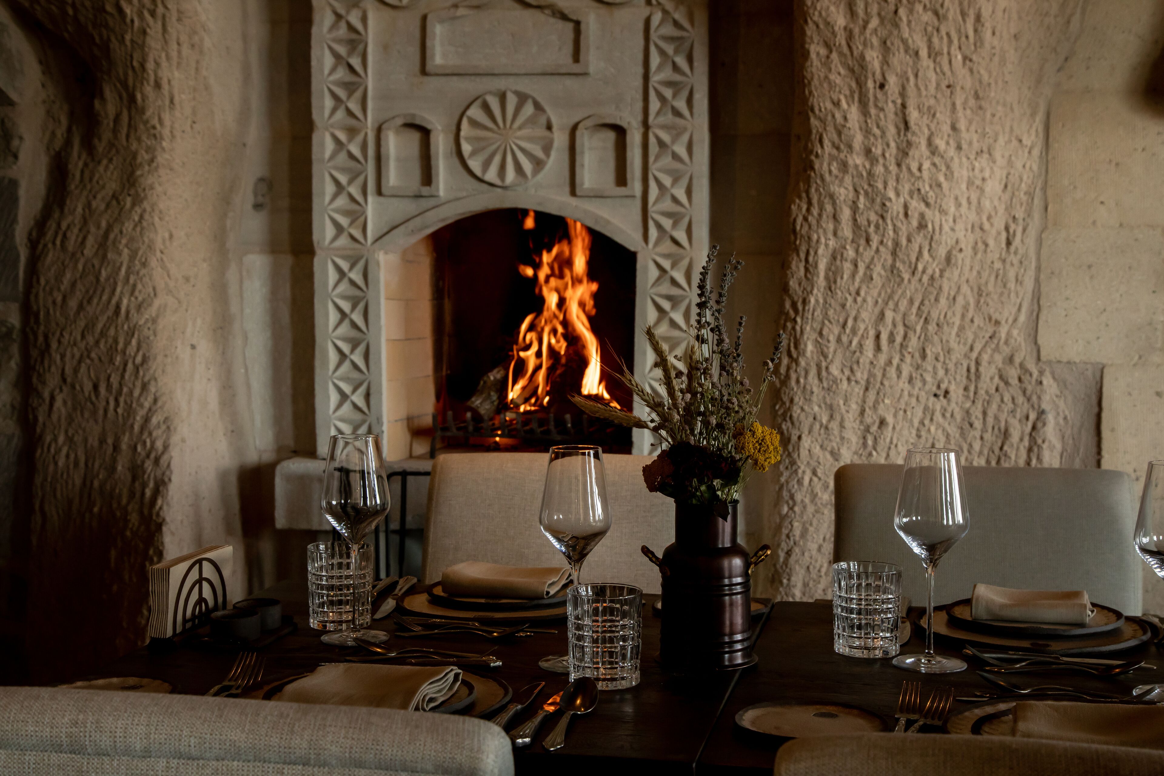 A dining table set with wine glasses, water glasses, and cutlery, with a fireplace in the background.