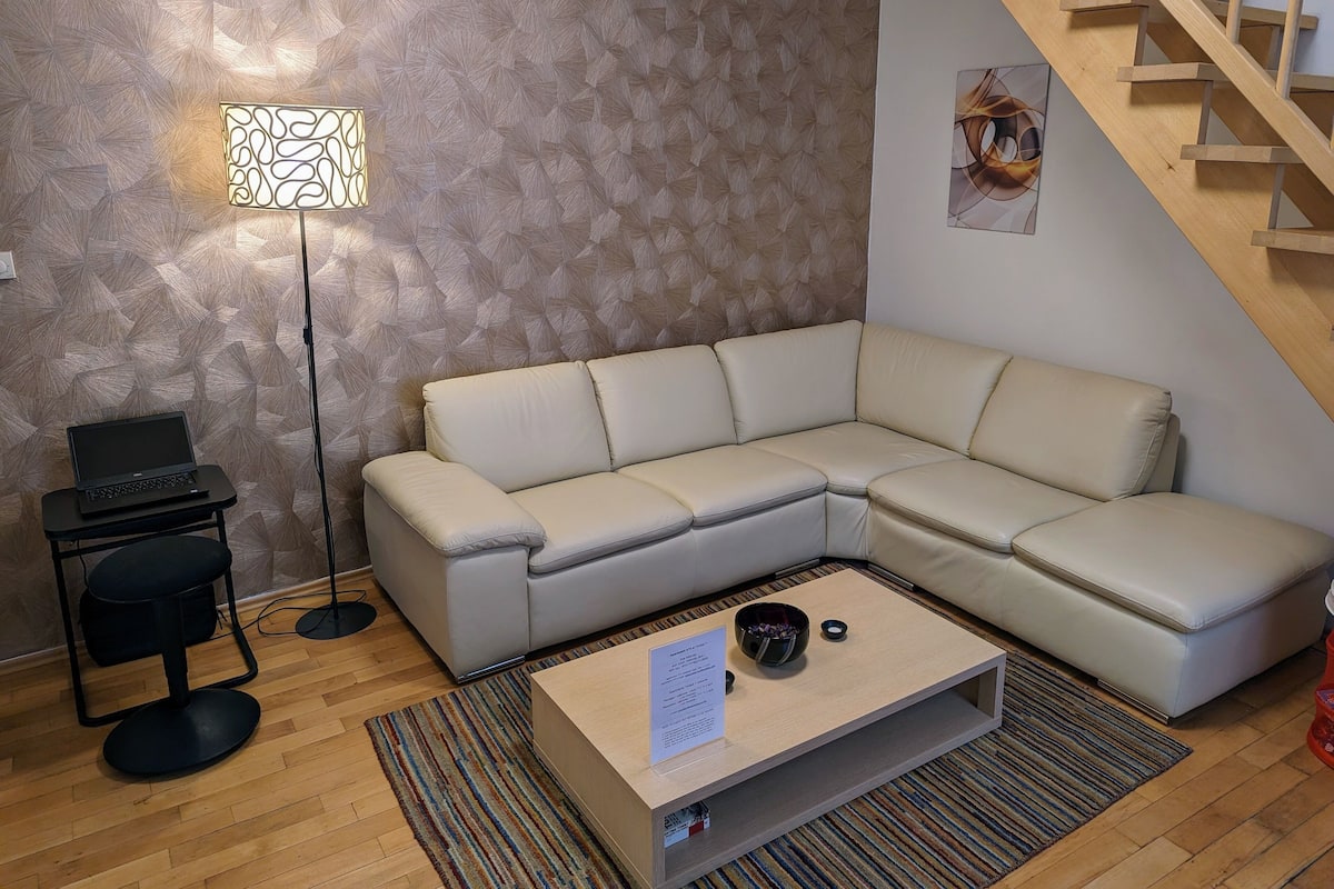 Un salon moderne avec un canapé d’angle beige, une petite table basse sur laquelle est posé un panneau, un lampadaire et un escalier.