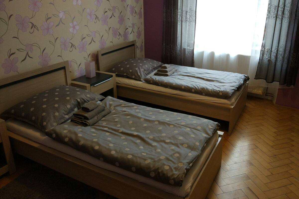 Une chambre avec deux lits simples, une table de chevet, un mur recouvert d’un papier peint à motifs floraux et une fenêtre avec des rideaux.