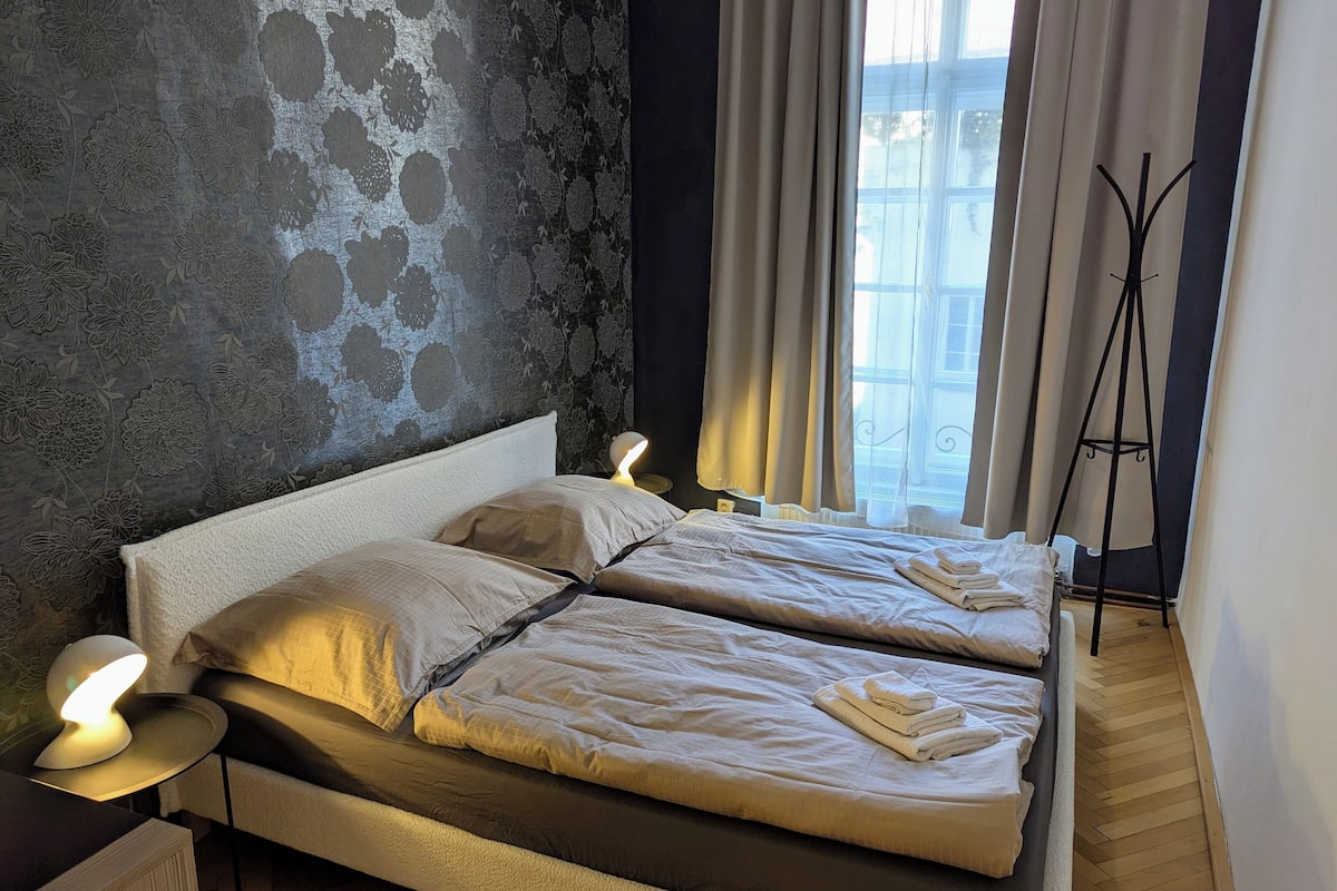 Un lit double avec du linge de lit blanc, une table de chevet avec une lampe, et une vue sur l’extérieur par une fenêtre avec des rideaux.