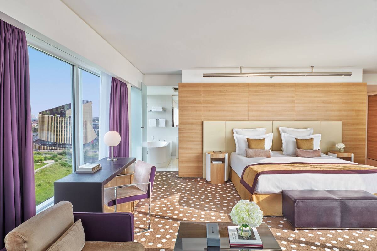 A modern hotel room with a large bed, a desk, a chair, and a view of the city.