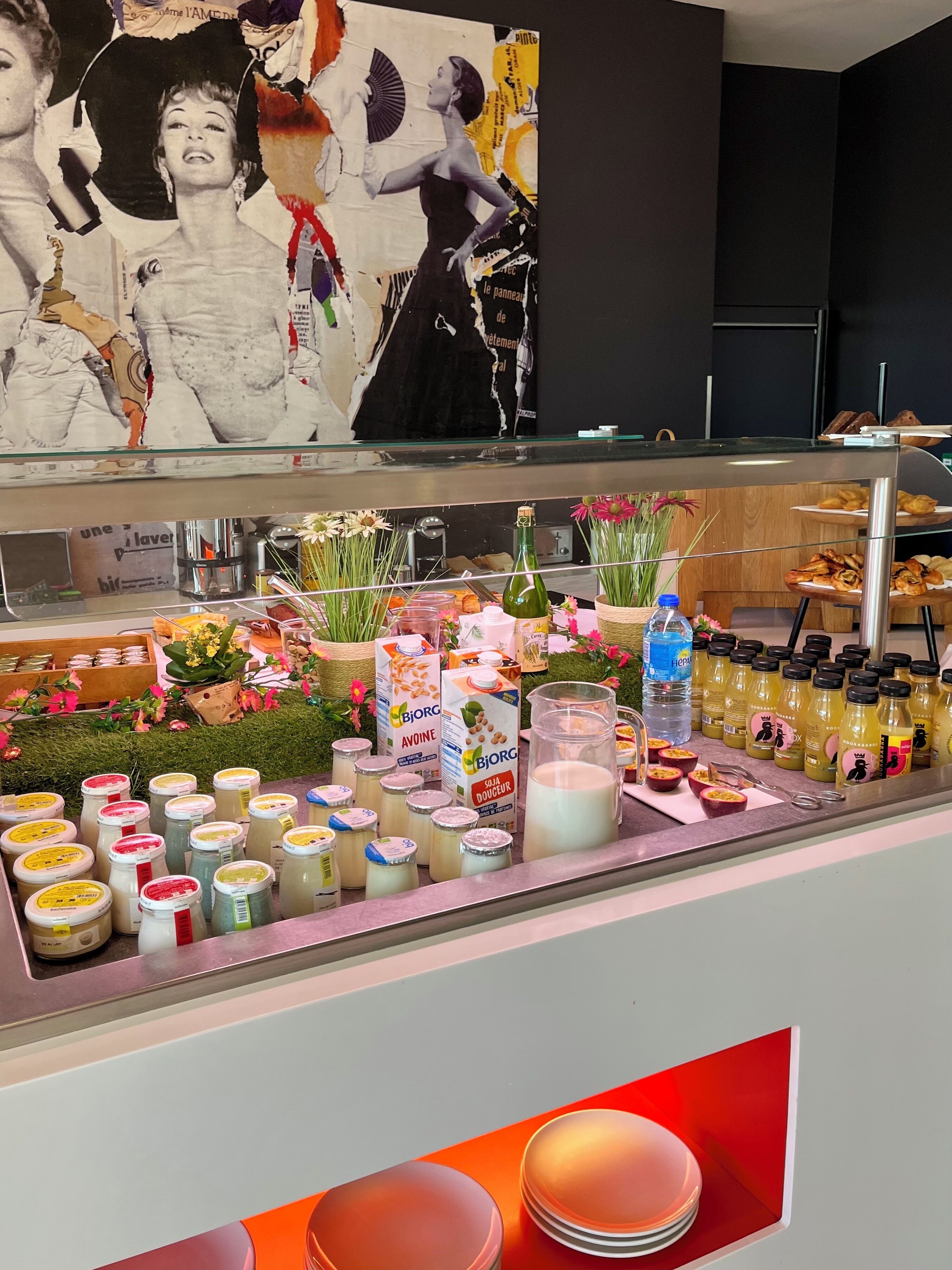 A counter with various food items and drinks, including jars of condiments, a bottle of olive oil, and fresh flowers.