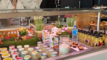 A counter with various food items and drinks, including jars of condiments, a bottle of olive oil, and fresh flowers.