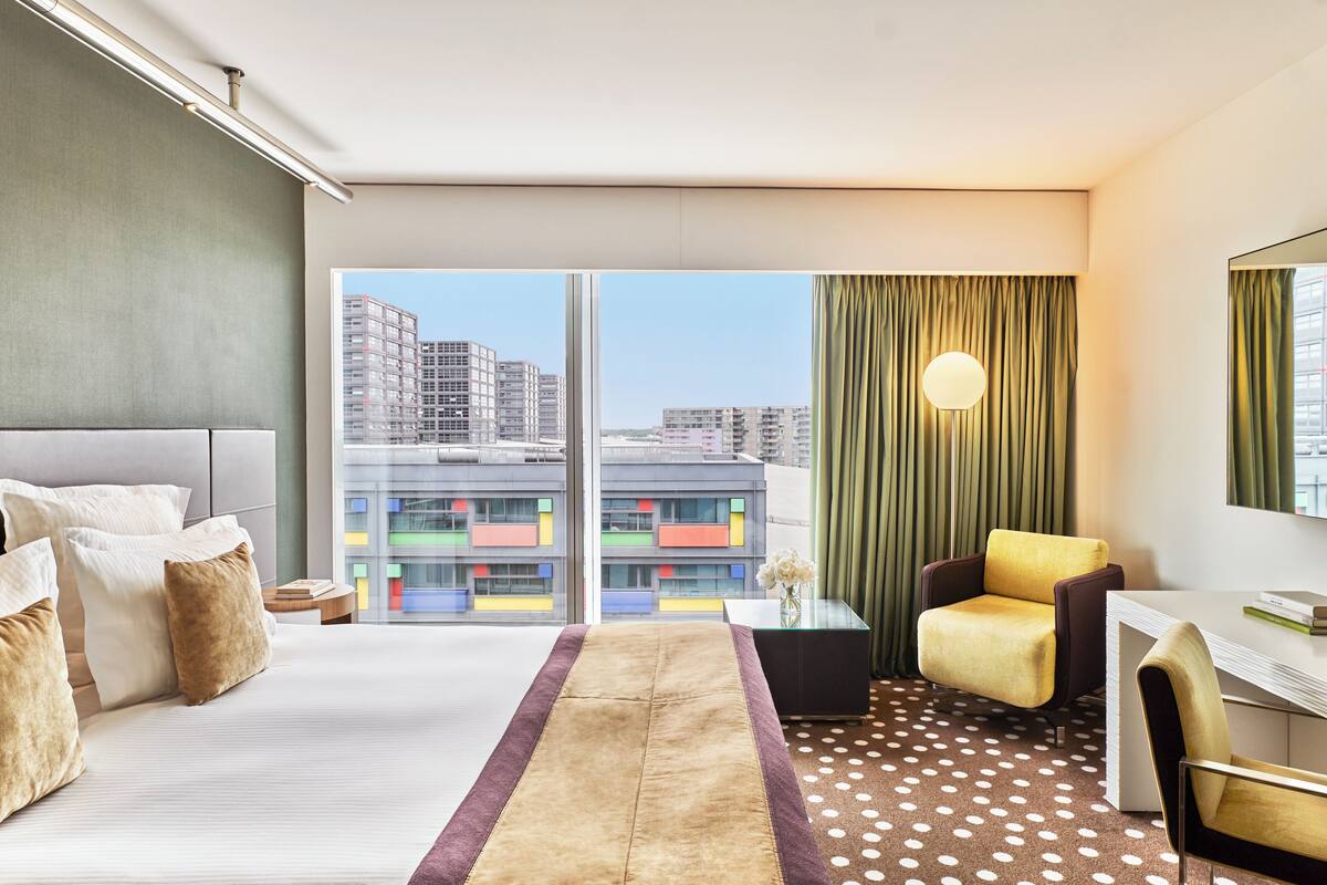A hotel room with a large bed, a desk, a chair, and a view of buildings through the window.