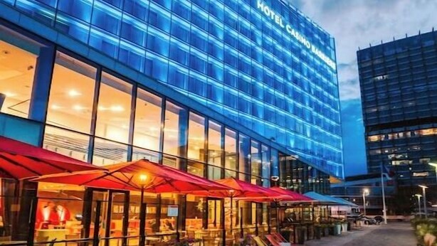 Un hotel moderno con fachada de vidrio, zona de estar al aire libre con sombrillas de colores y un sendero pavimentado.