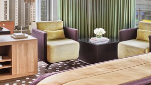 Habitación de hotel con una cama grande, dos sillones, una mesita con un florero y vistas a un paisaje urbano a través de amplios ventanales.