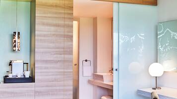 A modern hotel room with a bed, a desk with a chair, a vanity with a mirror, and a bathroom visible through an open door.