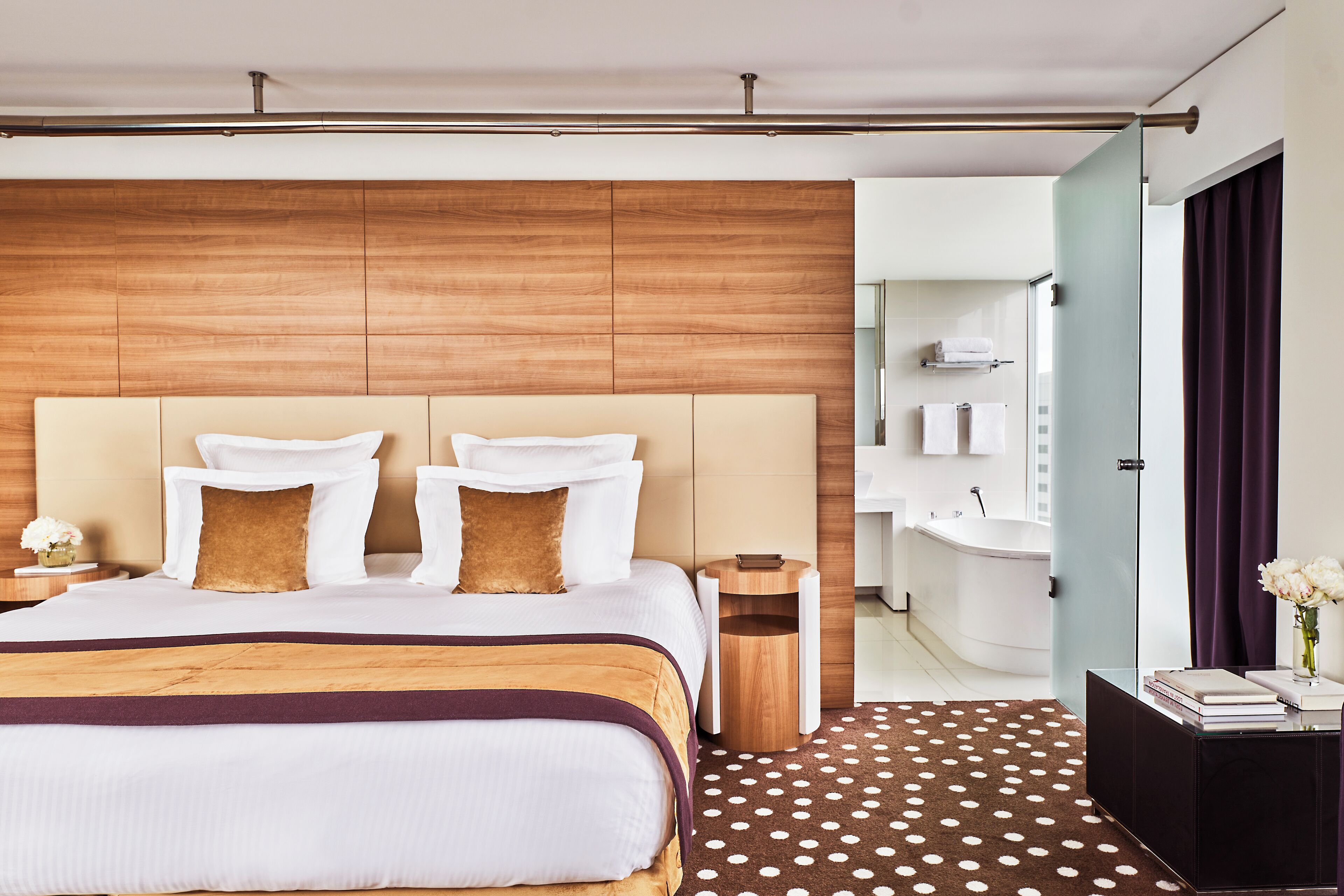 Una habitación de hotel moderna con una cama grande, cabecera de madera, una mesita de noche pequeña, una mesilla auxiliar y un baño con bañera, además de un tapete estampado de lunares.