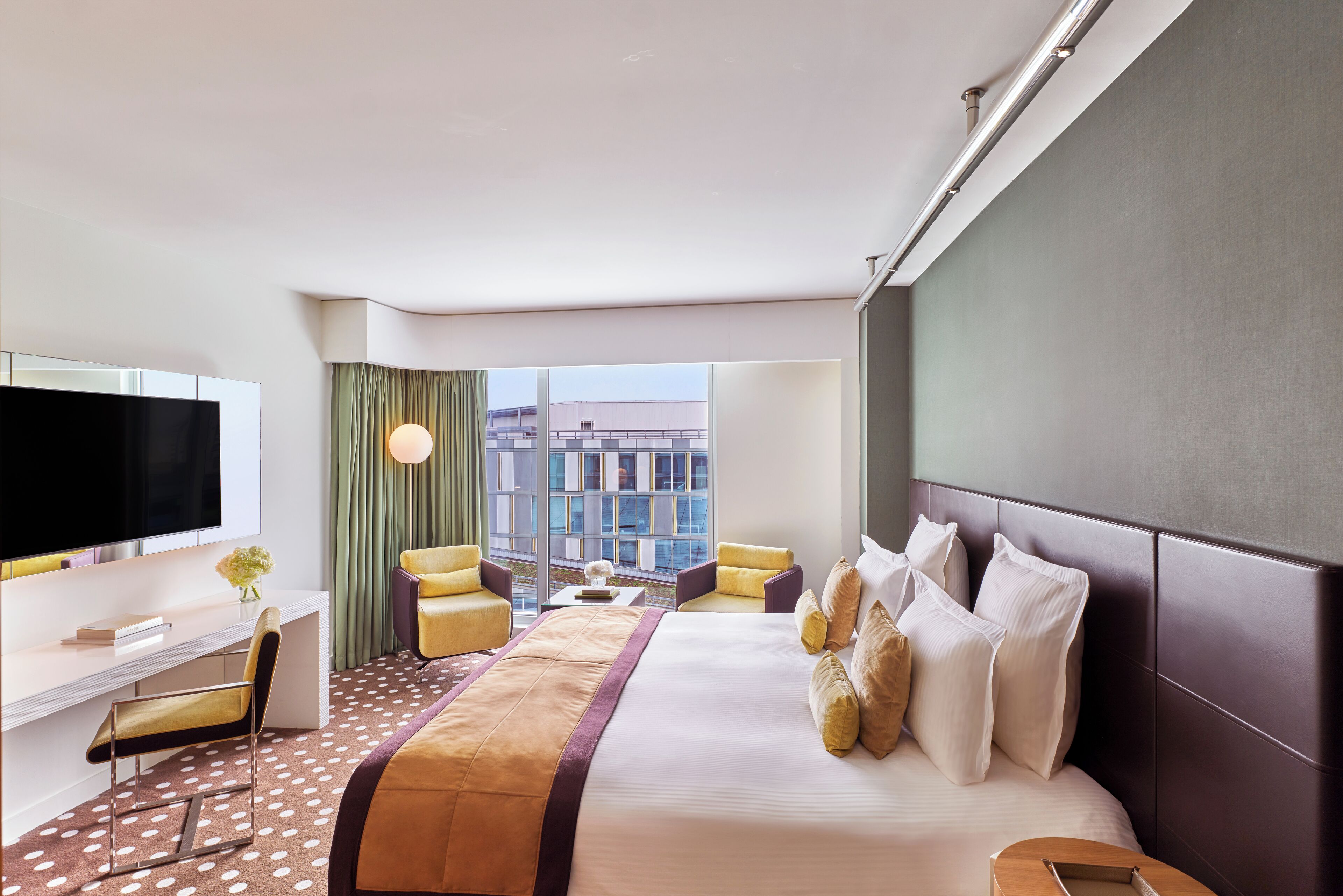 A modern hotel room with a large bed, two armchairs, a desk with a flat-screen TV, and a view of the cityscape through large windows.