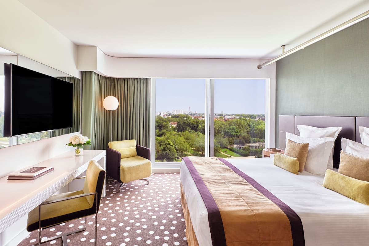 A hotel room with a large bed, a desk, a chair, a TV, and a view of a cityscape.