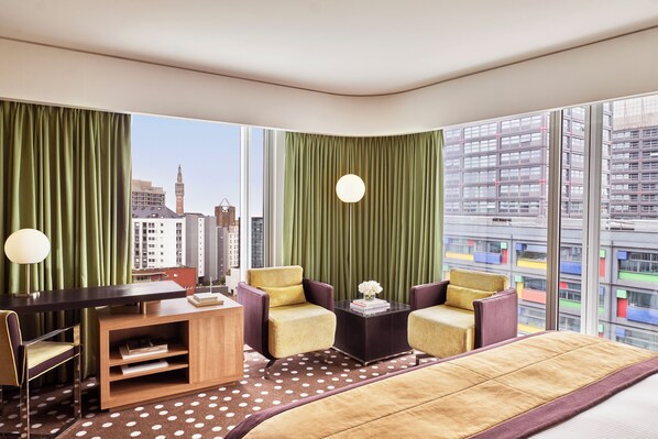 Una habitación de hotel moderna con una cama grande, dos sillones, un escritorio y vistas a la ciudad.