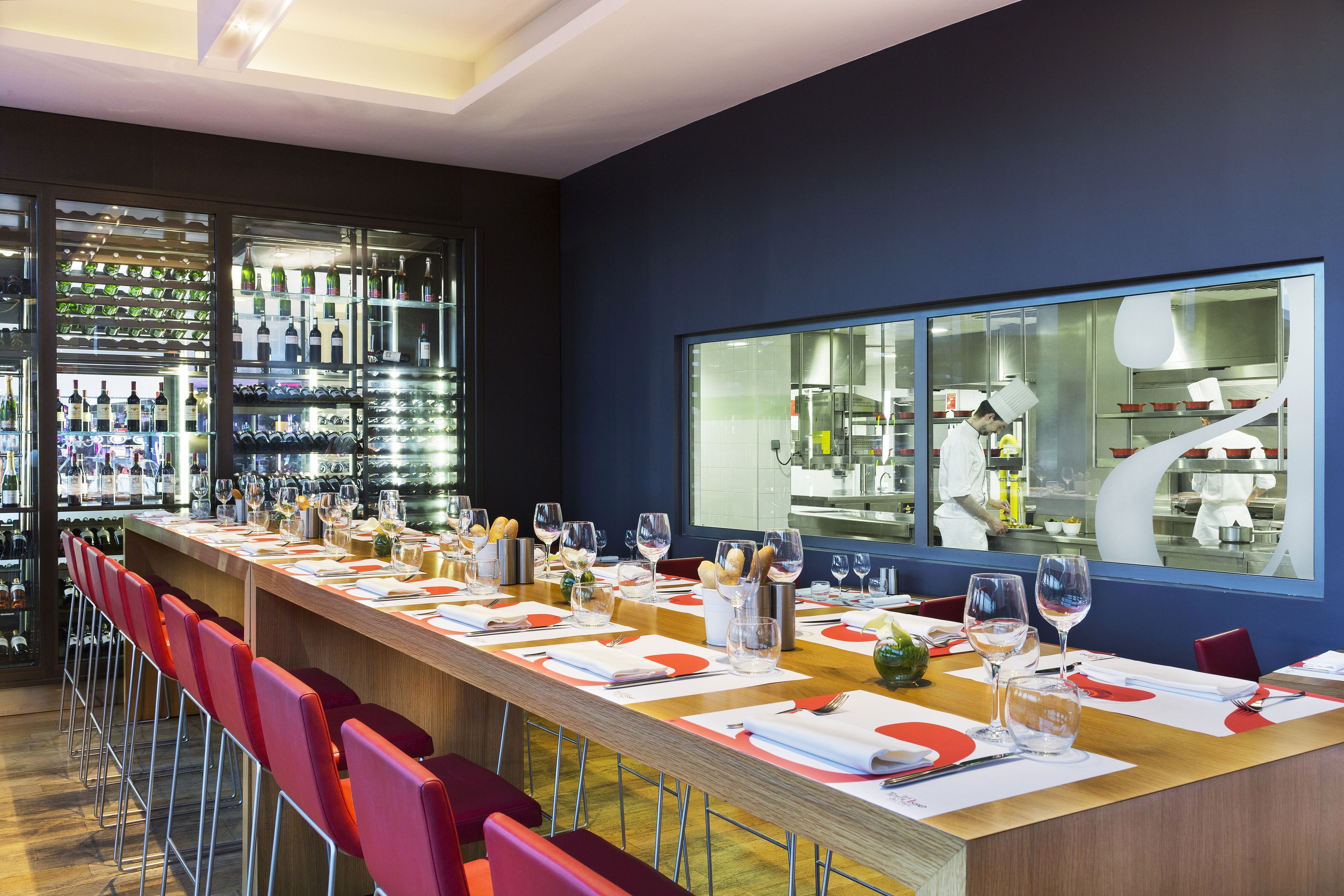 A restaurant with a long counter, red chairs, and a wine rack in the background.