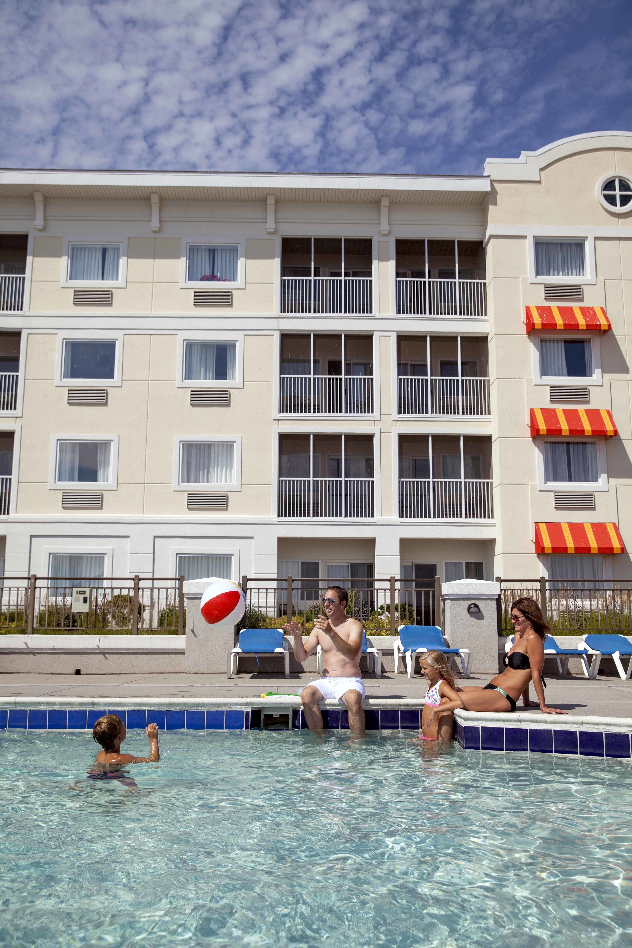 A hotel pool area with clear blue water, lounge chairs, and a building with balconies and windows.