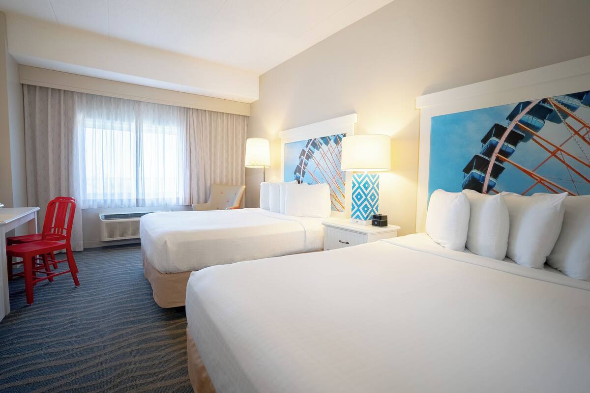 Habitación de hotel con dos camas, un escritorio, una silla y un amplio ventanal con cortinas.