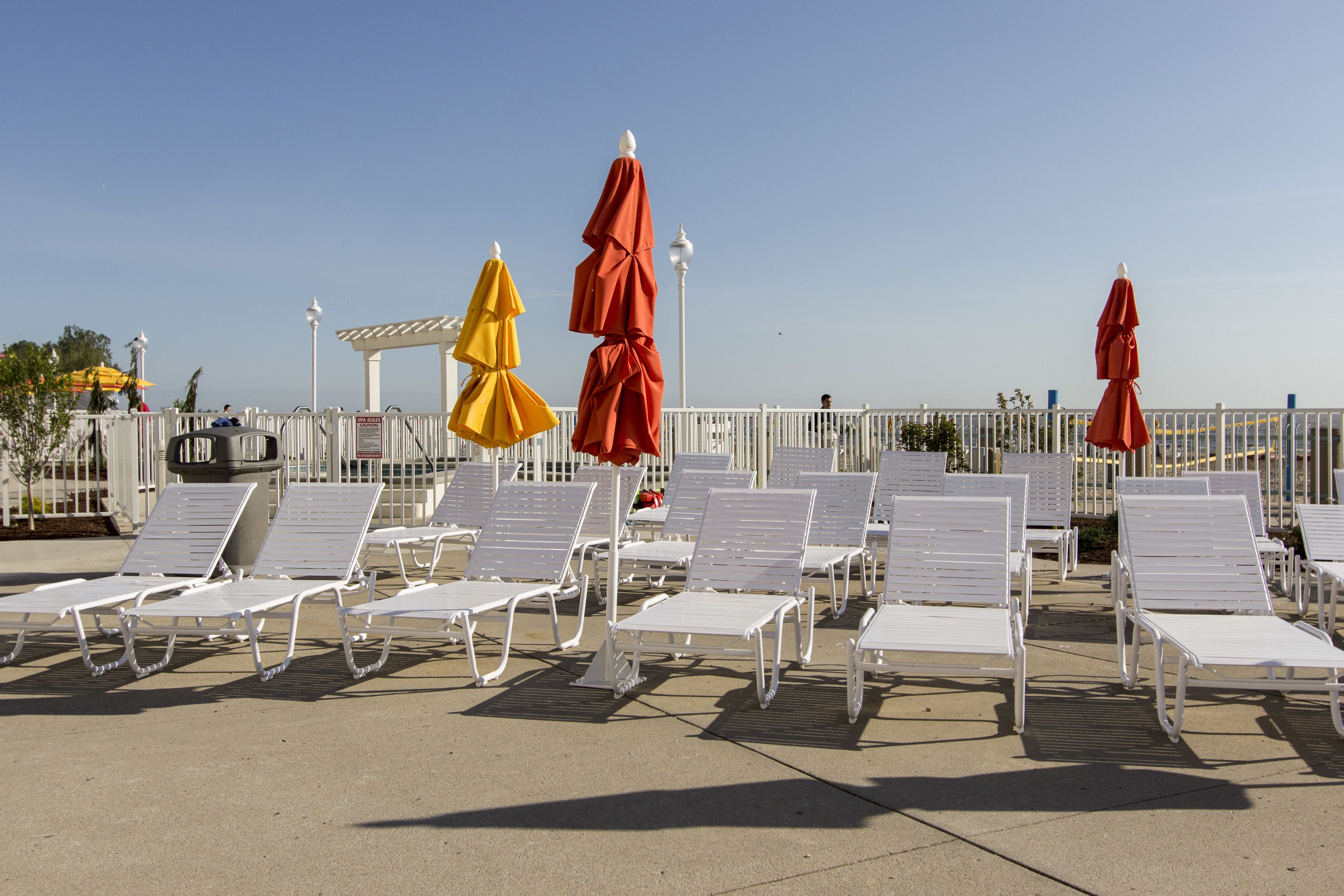 A row of white lounge chairs and red umbrellas on a sunny terrace with a clear view of the sea.