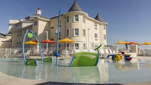 Un área de piscina con sillones de descanso y sombrillas, un edificio grande con torreón y un cielo azul despejado.