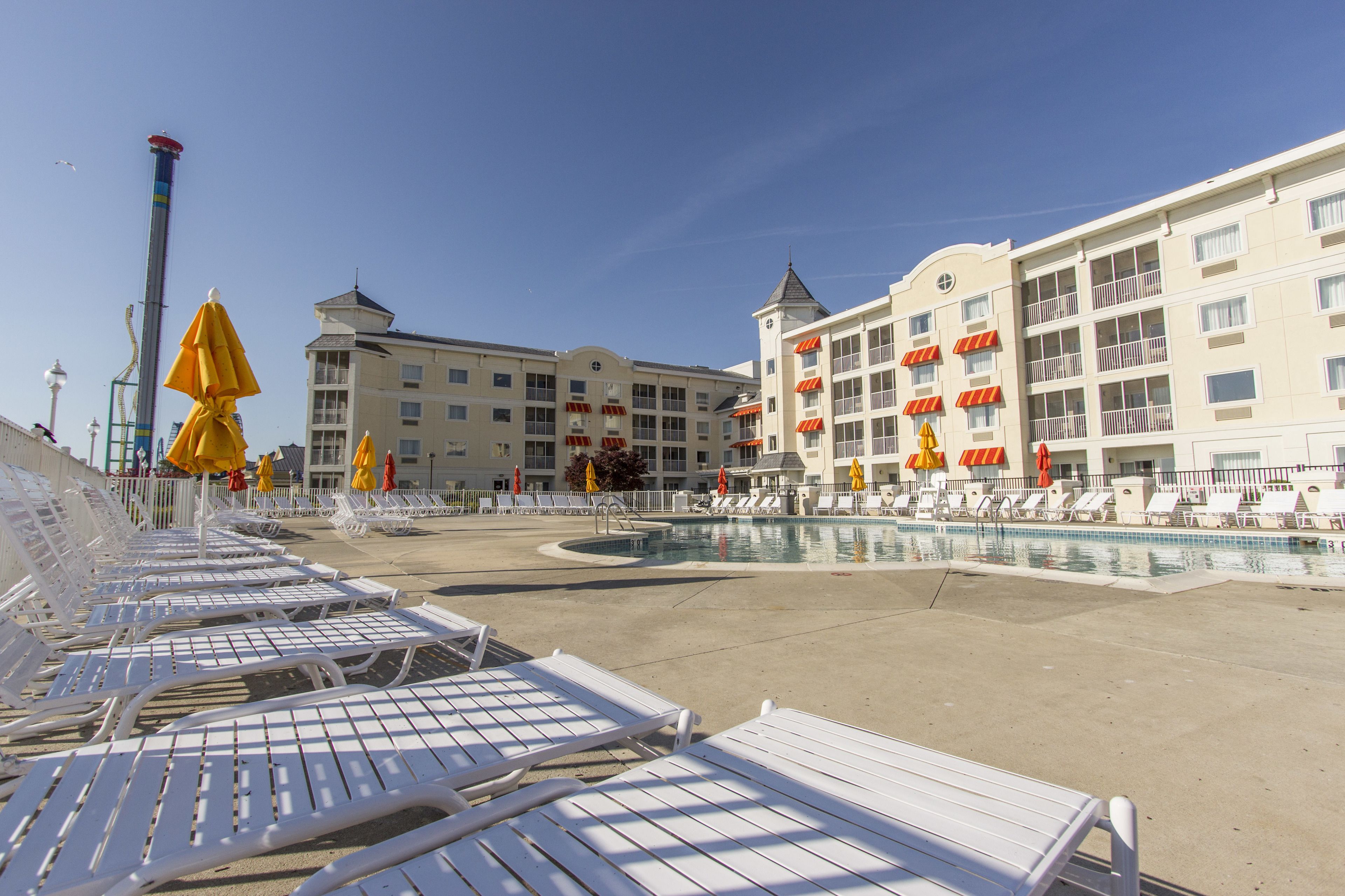 Une piscine d’hôtel entourée de chaises longues blanches et de parasols jaunes, sous un ciel dégagé, avec un bâtiment doté de nombreux balcons.