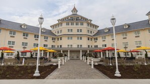 Un hotel grande de varios pisos con una entrada central, asientos exteriores con sombrillas y una fachada simétrica.