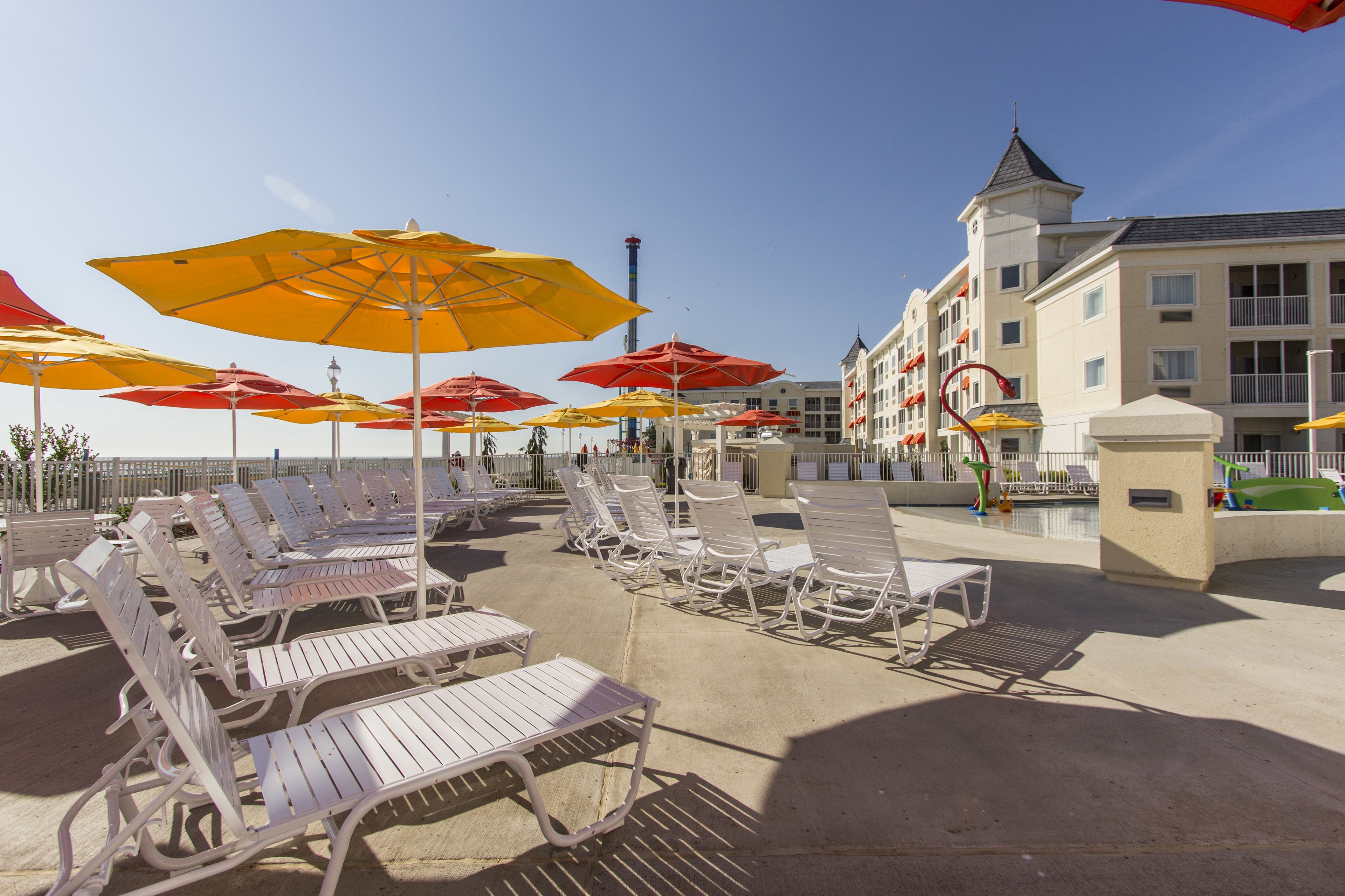 A beachside hotel with rows of white lounge chairs and colorful umbrellas, a pool, and a building with a tower.
