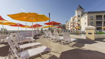 A beachside hotel with rows of white lounge chairs and colorful umbrellas, a pool, and a building with a tower.