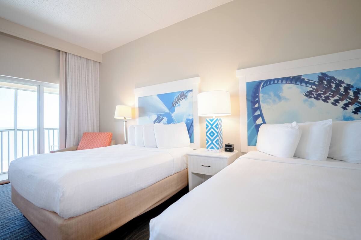 Una habitación de hotel con dos camas, una mesita de noche, una lámpara y un ventanal con cortinas.