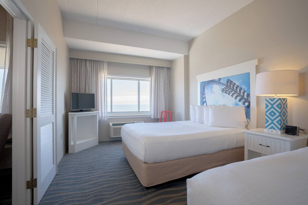 Una habitación de hotel con cama, escritorio con televisor, ventana con cortinas y un cuadro en la pared.